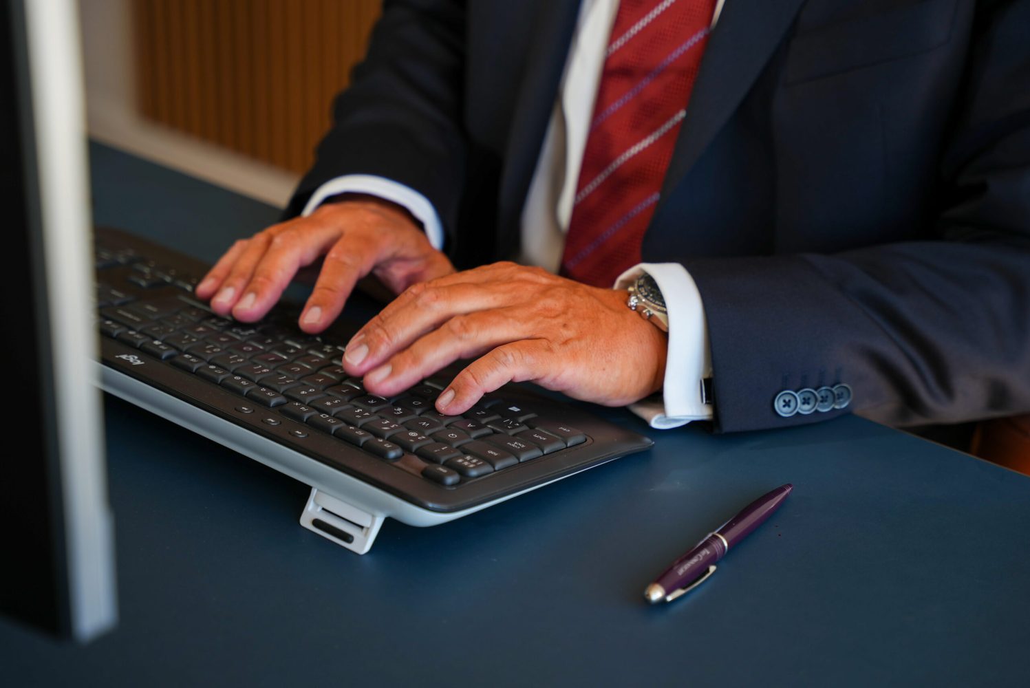 To mandehænder, der skriver på et tastatur. Ved siden af tastaturet ligger der en kuglepind